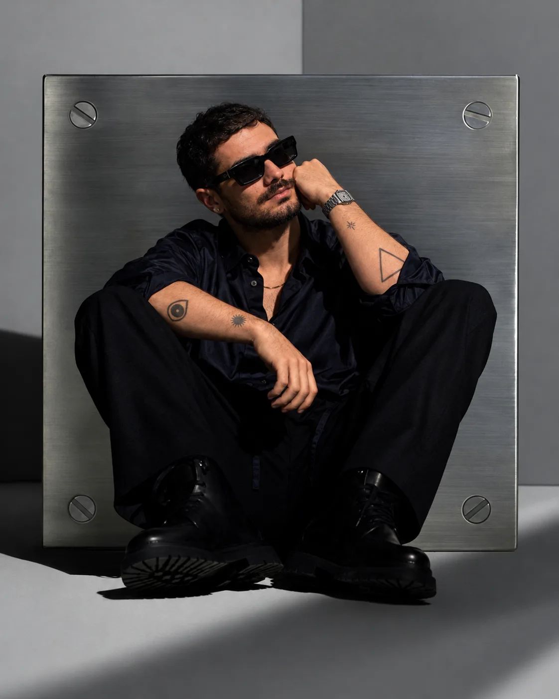 Ray Ramzi in a black shirt and sunglasses, sitting in front of a steel panel