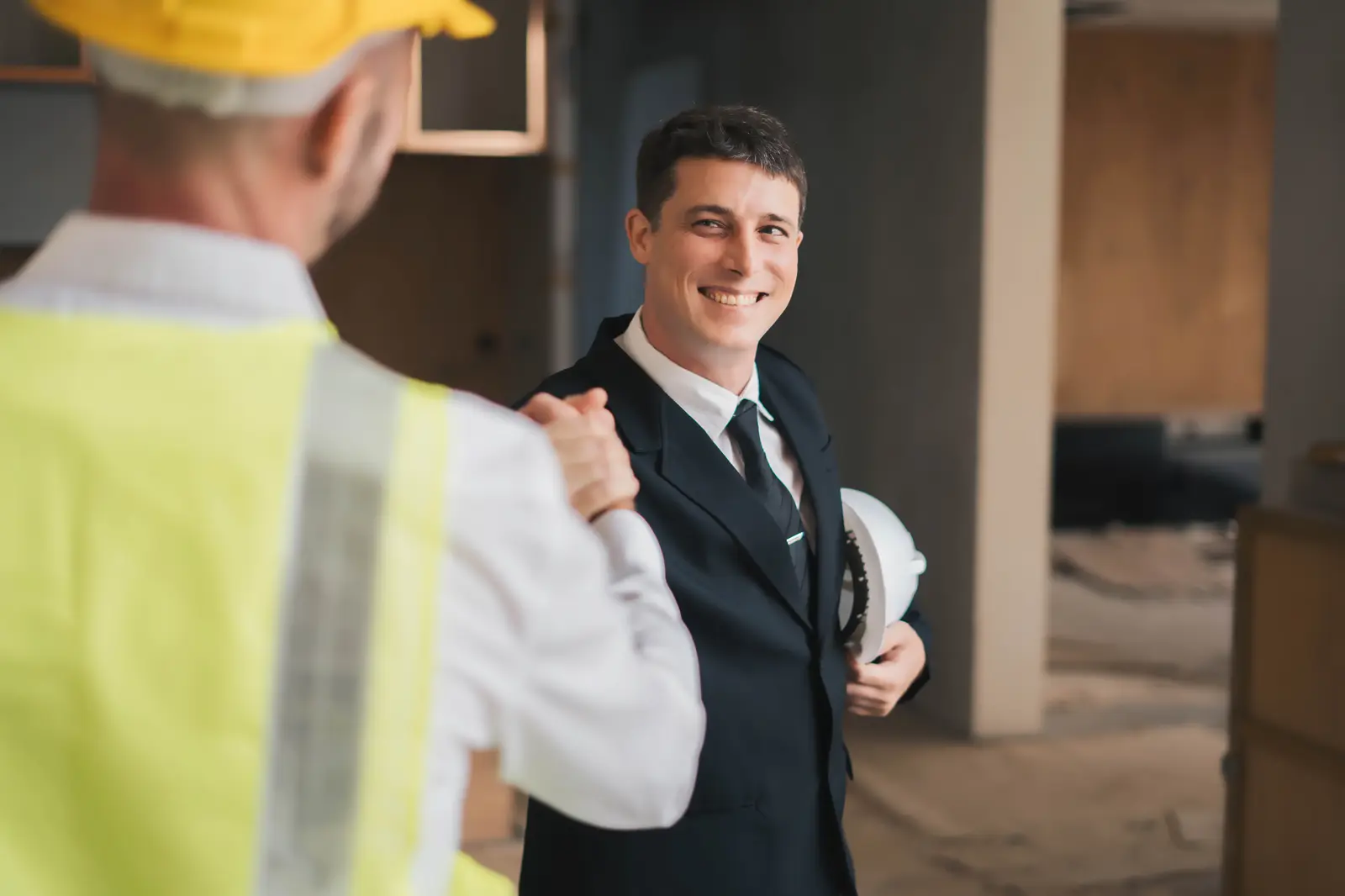 A smiling agent in a suit, holding a hard hat, shaking hands with a contractor in a safety vest