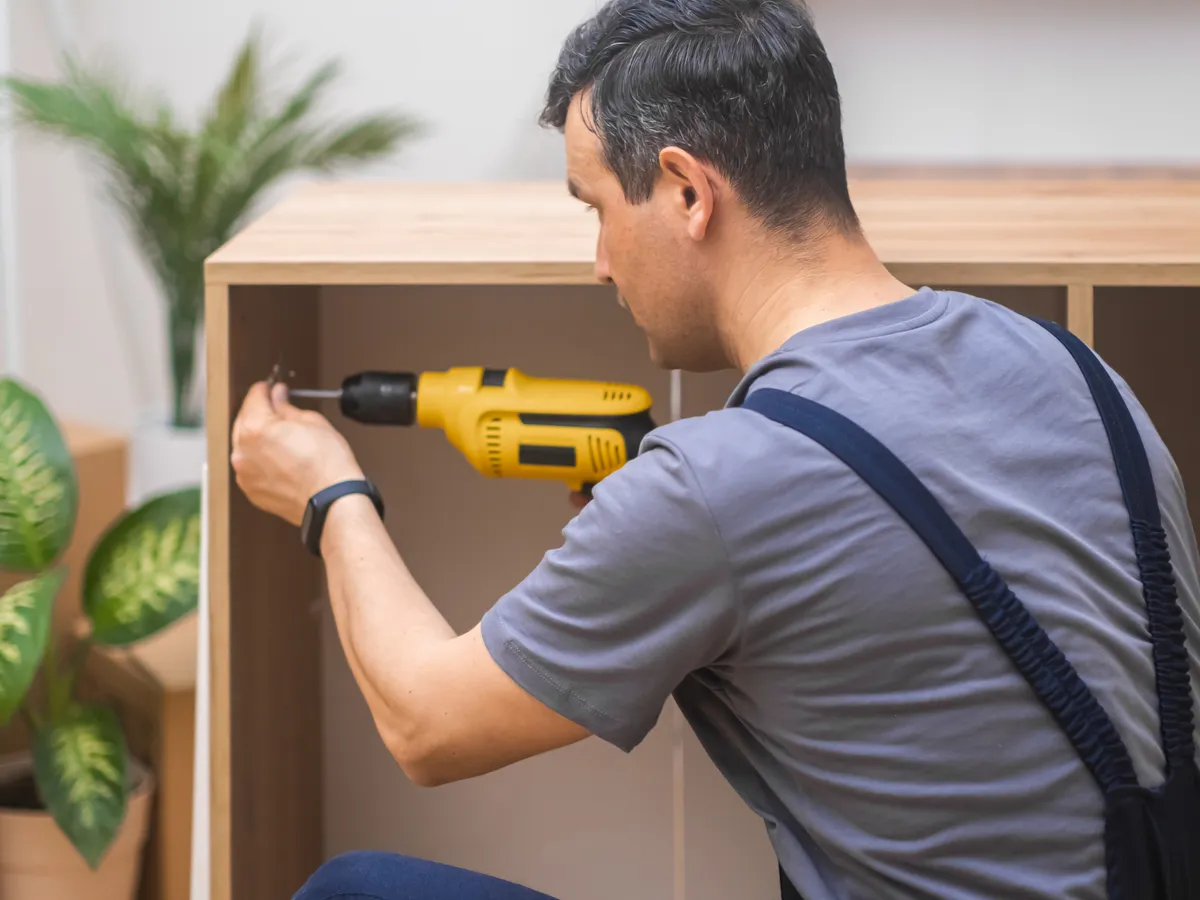 Handyman with a yellow drill assembling a wooden cabinet in a living room