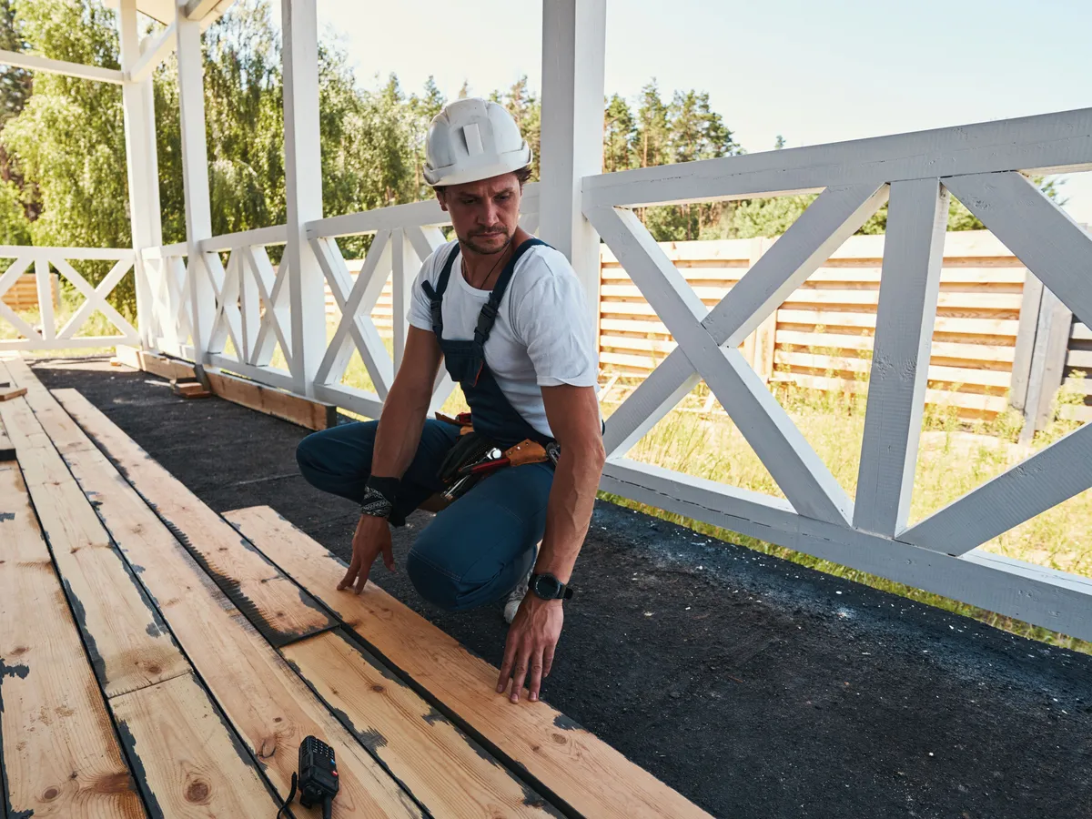 Carpenter in a hard hat laying new boards on a porch with a white railing