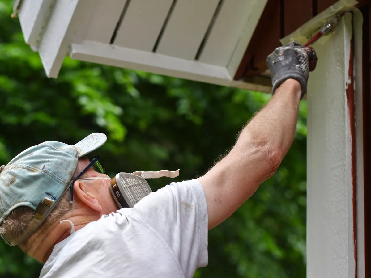 Painter in a respirator brushing fresh paint onto exterior trim under the eaves