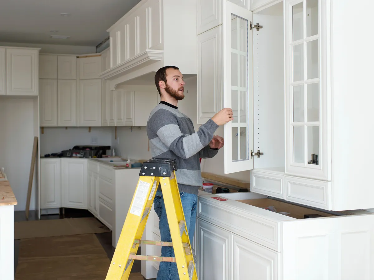 Craftsman on a stepladder adjusting a glass-panel cabinet door in a white kitchen