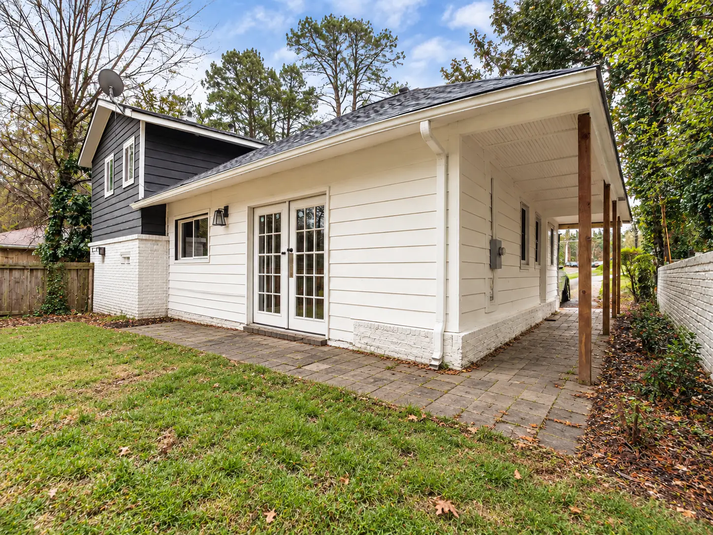 Back of a white cottage with French doors and a paver patio running along the house and under a covered side walk, beside a grass lawn