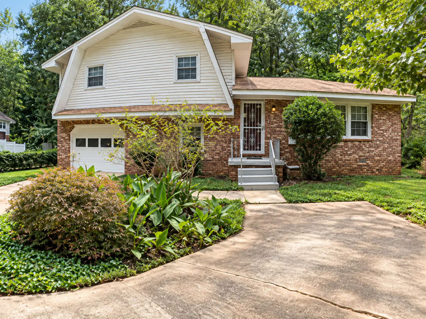 Split-level house before renovation, with red brick, beige vinyl siding, a brown roof, a white security door and an overgrown front bed