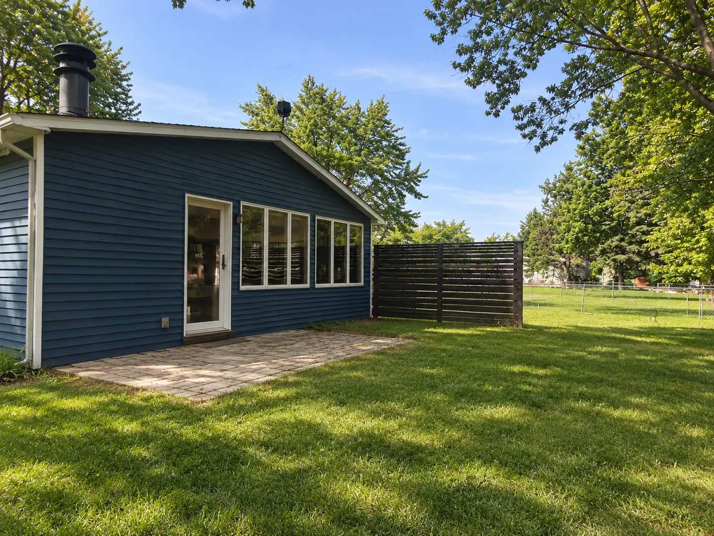 Back of a blue-sided house with a glass door, a small paver patio, a dark slatted privacy screen and a large lawn