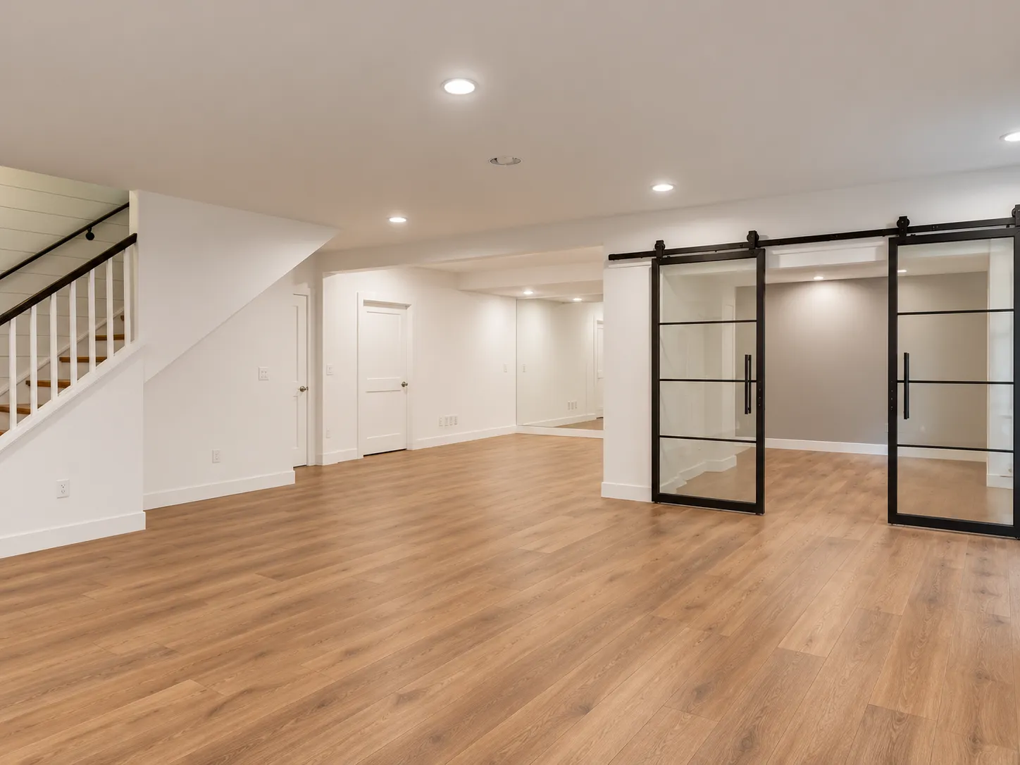 The same basement finished, with white walls, recessed lights, wood-look plank floors and black-framed glass barn doors