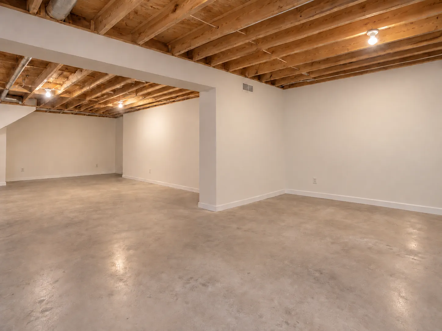 Unfinished basement before renovation, with exposed wood joists and ductwork, white painted walls, bare bulbs and a concrete floor