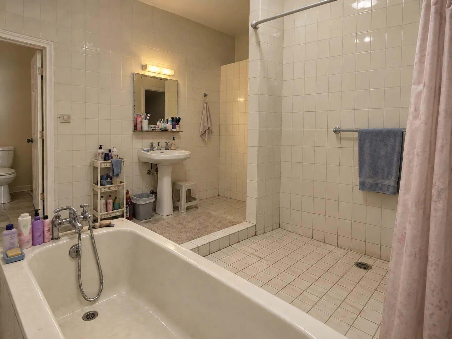 Bathroom before renovation, with cream square tile on every wall, a tiled-in tub, a pedestal sink and an open shower behind a pink curtain
