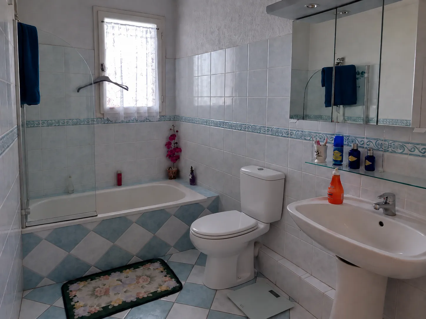 Small bathroom before renovation, with pale blue tile, a decorative border, a tub with a glass screen and a pedestal sink