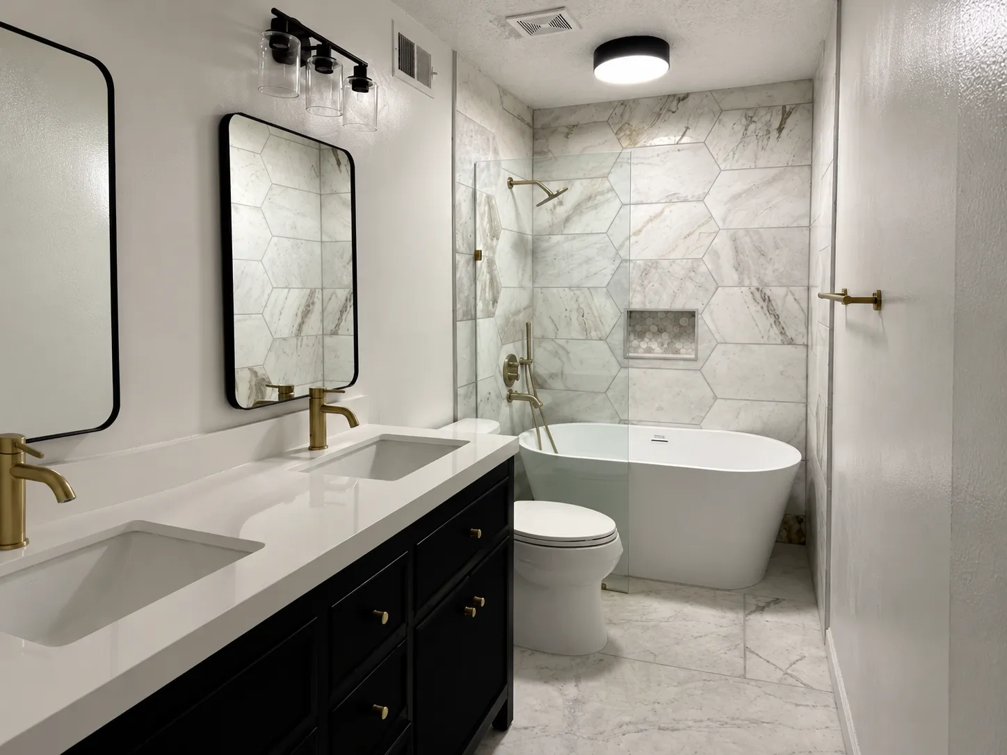The same bathroom after renovation, with a black double vanity, brushed-gold faucets, a freestanding tub and a wall of hexagon marble-look tile