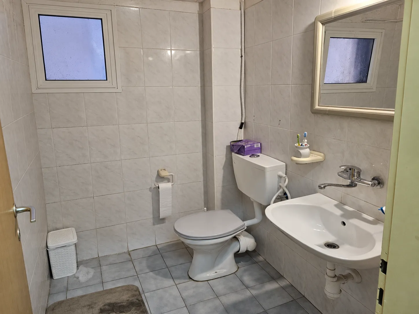 Powder room before renovation, with grey tile on every wall, a wall-hung basin and exposed hoses behind the toilet
