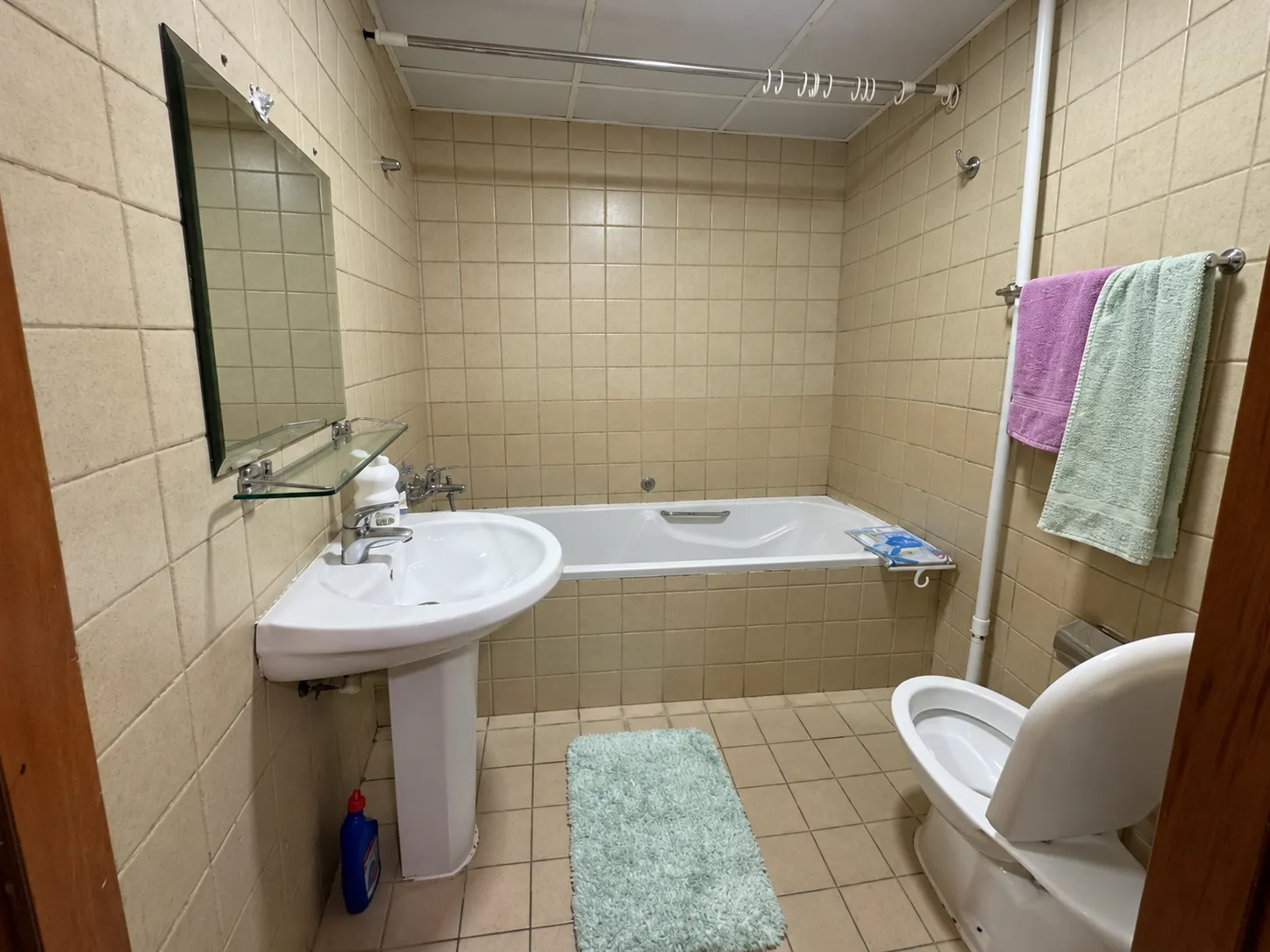 Bathroom before renovation, with speckled beige tile on every wall, a drop-in tub and a pedestal sink