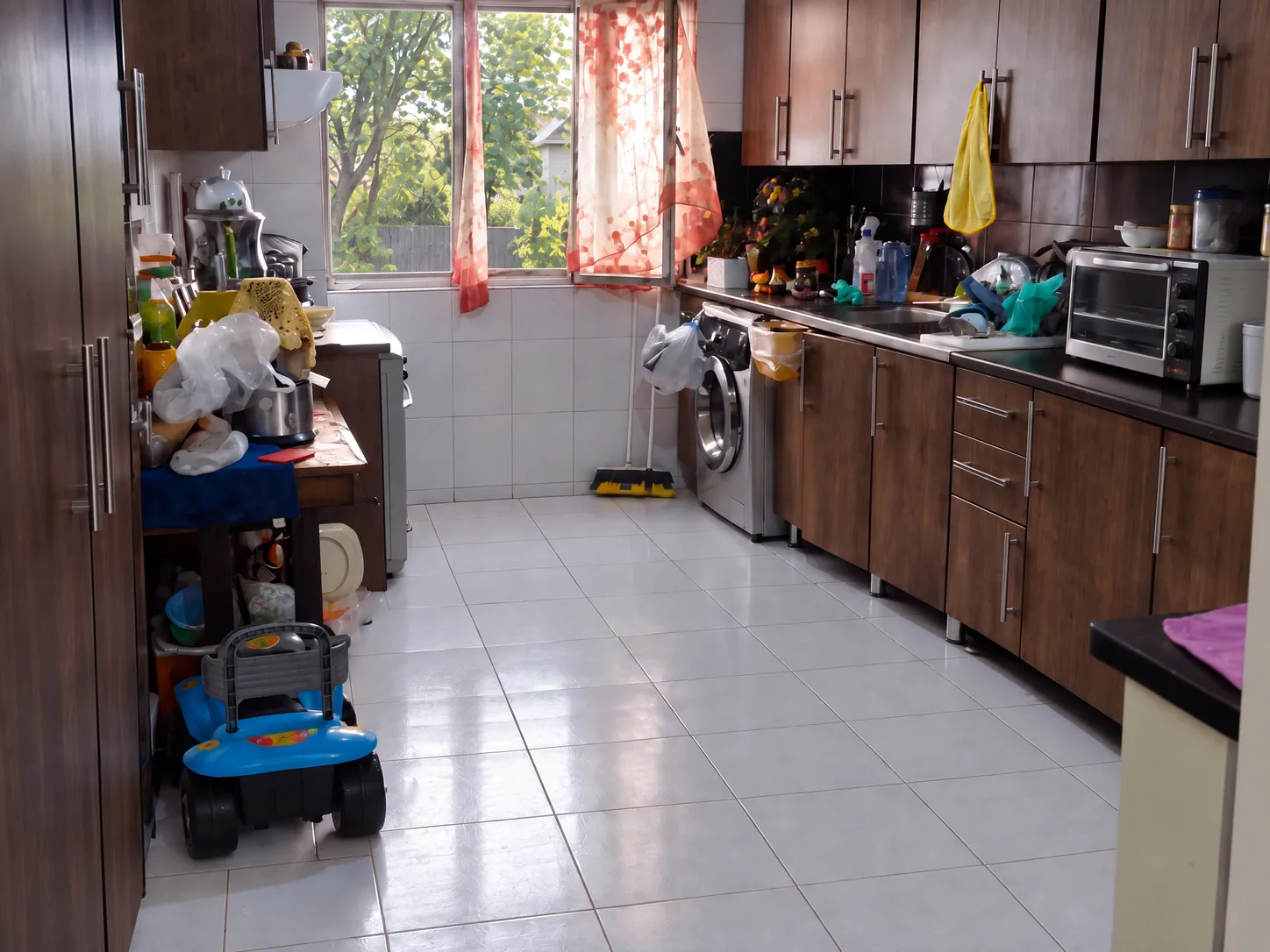 Cluttered galley kitchen before renovation, with brown laminate cabinets, black countertops, white wall tile and a washing machine in the cabinet run