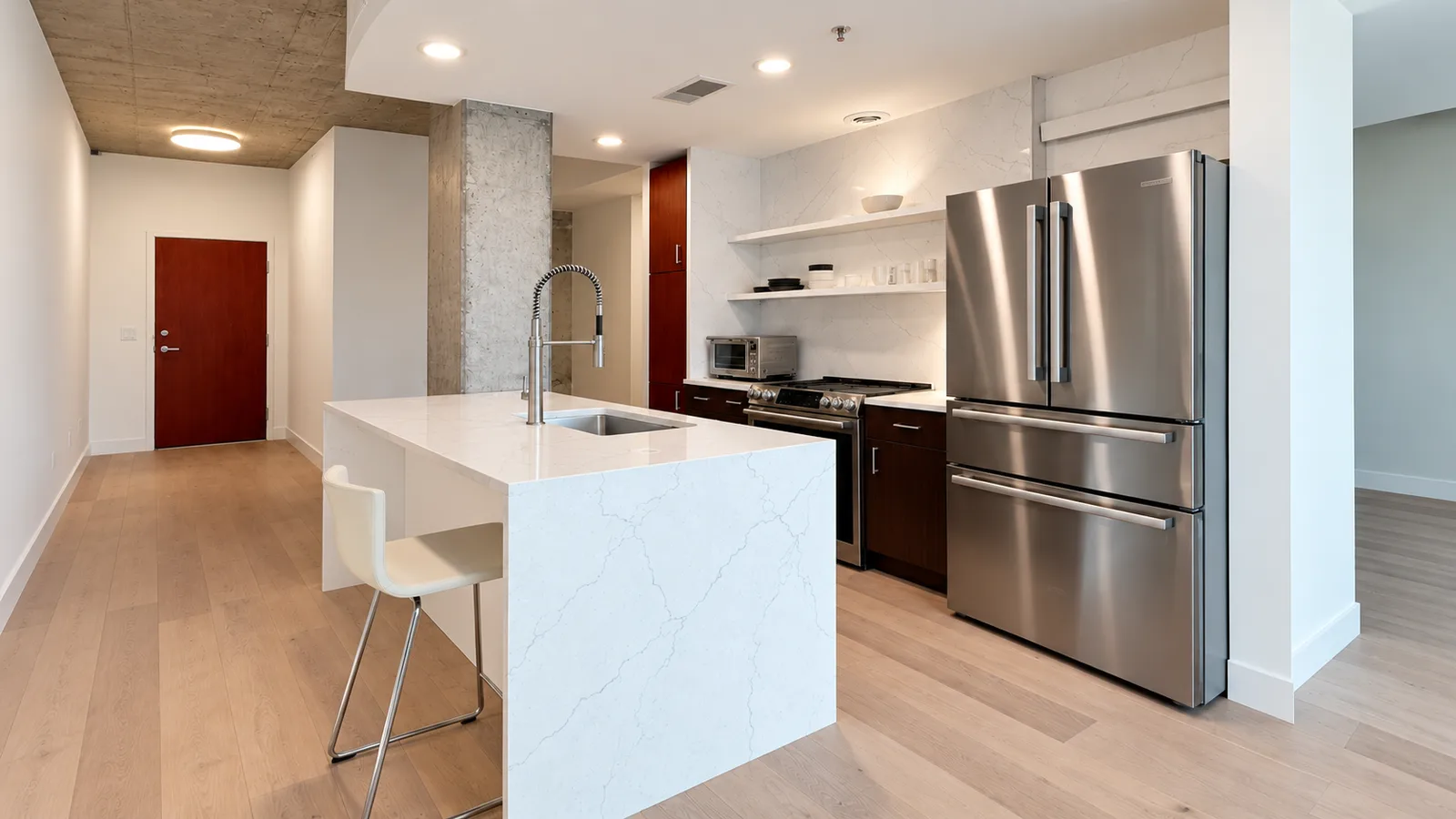 The same loft kitchen after renovation, with a white waterfall quartz island, quartz backsplash and shelves, and dark slab cabinets