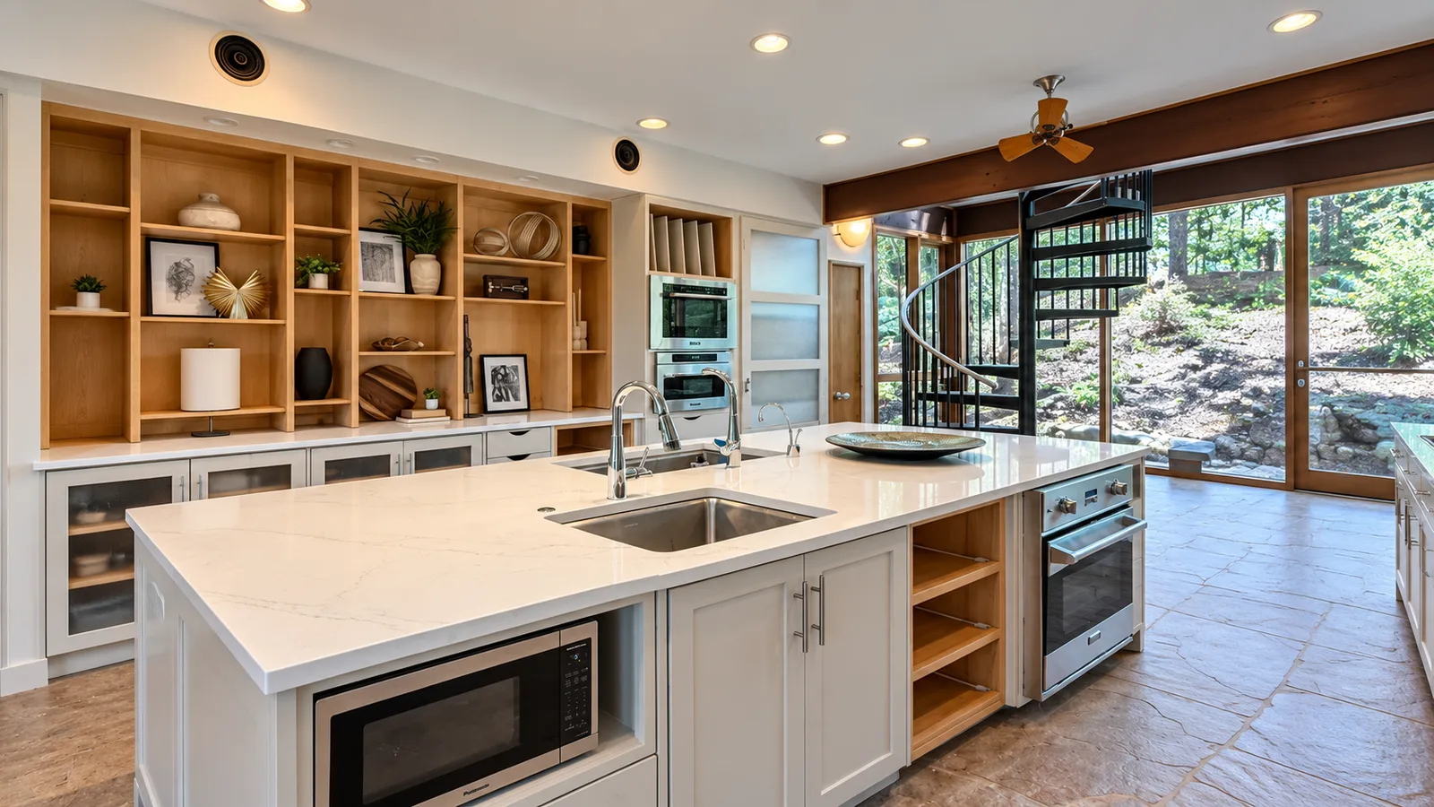 The same kitchen after renovation, with a wall of maple open shelving, a large white quartz island with two sinks and stainless built-in ovens