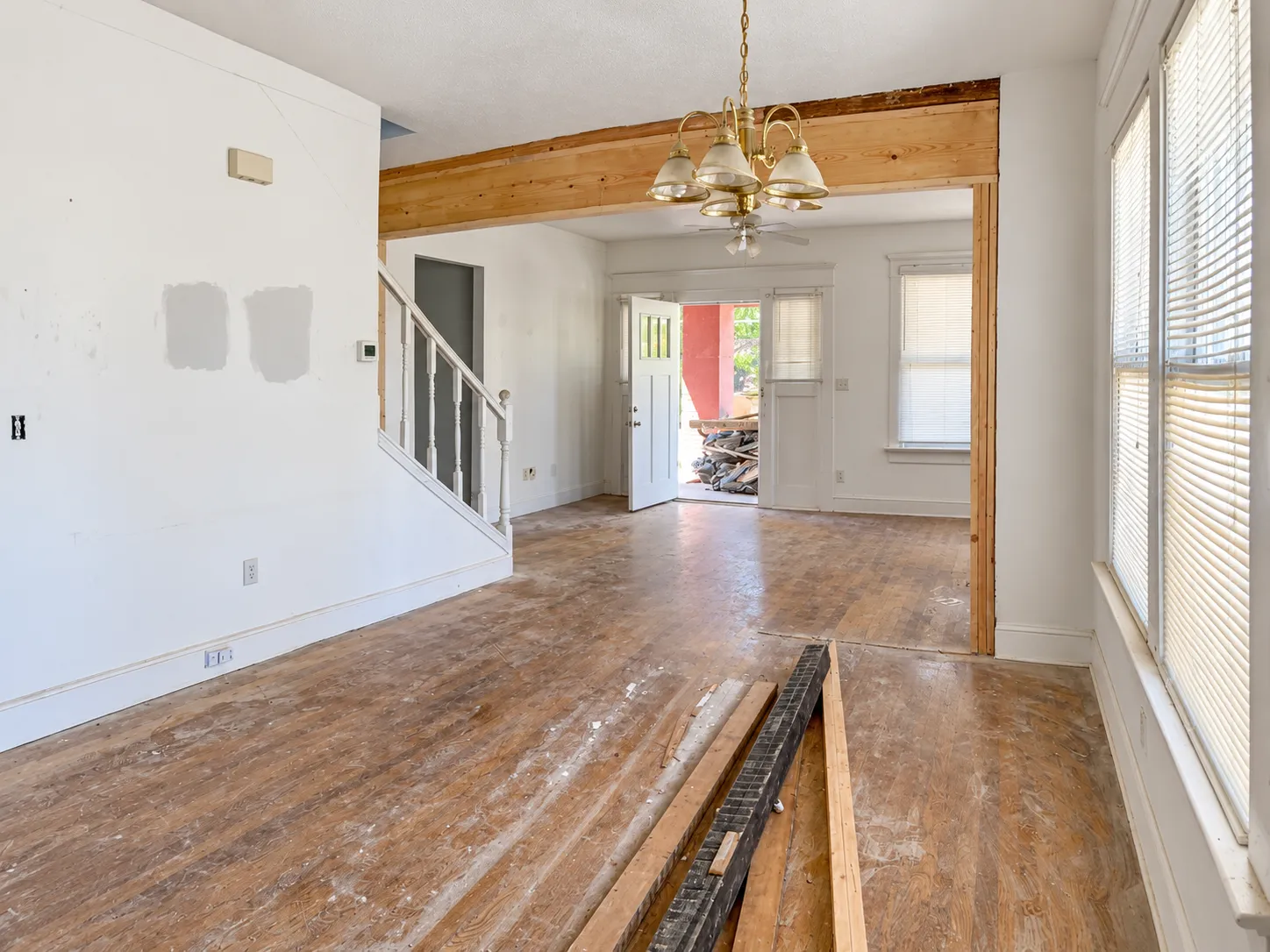 Living room mid-renovation, with scuffed wood floors, paint patches, lumber on the floor and a raw wood beam across the opening to the dining room