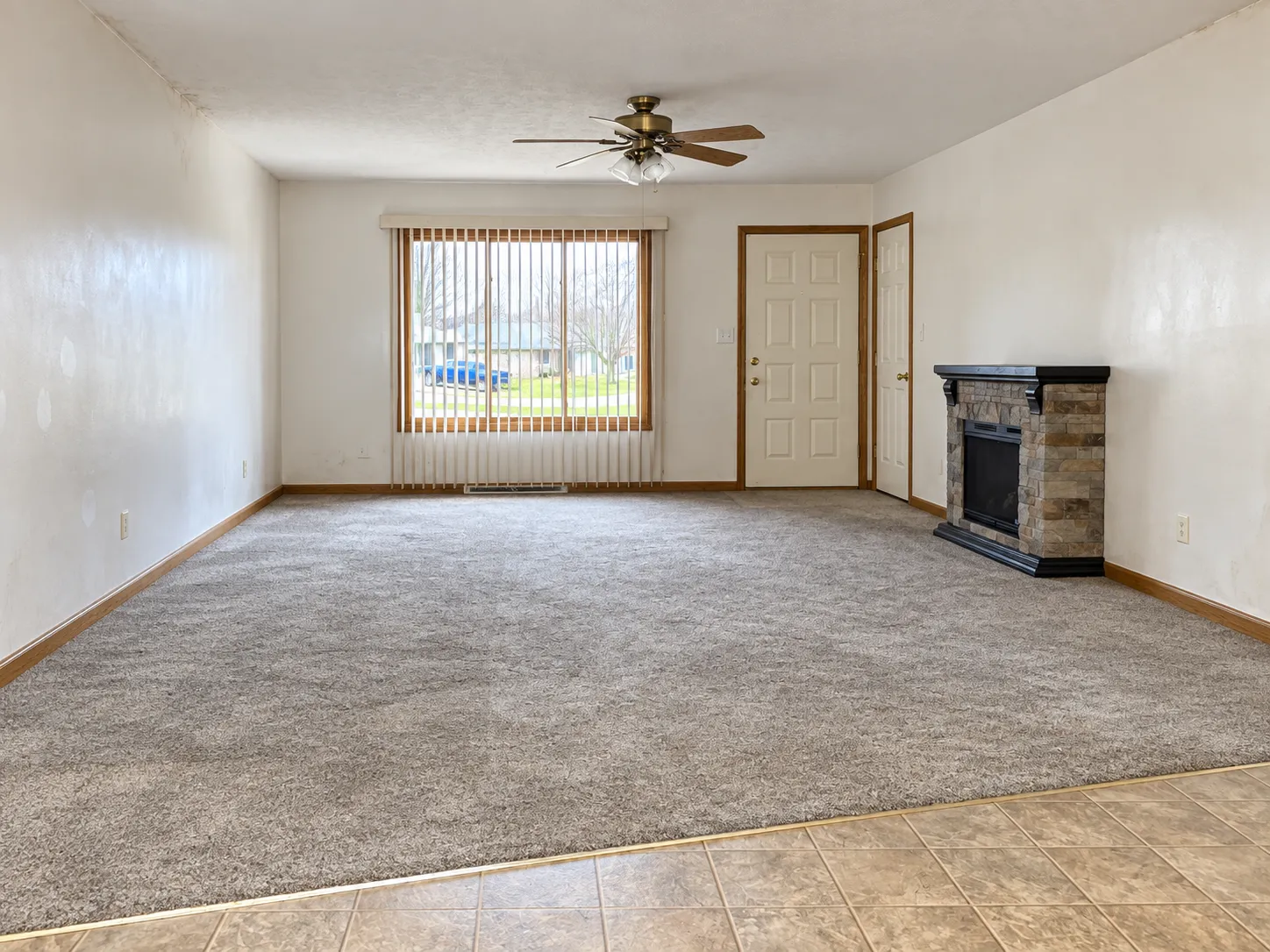 Living room before renovation, with grey carpet, a freestanding electric fireplace, vertical blinds and white walls with oak trim
